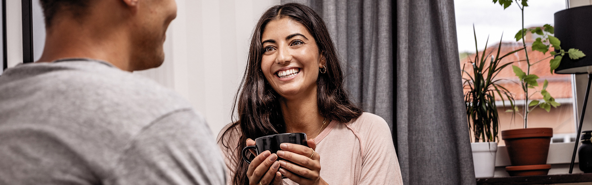 Två personer sitter vid ett köksbord och samtalar. En kvinna ler och håller en kaffekopp. En man sitter mittemot med ryggen mot kameran. I bakgrunden syns ett fönster med gardiner och flera krukväxter på fönsterbrädan.
