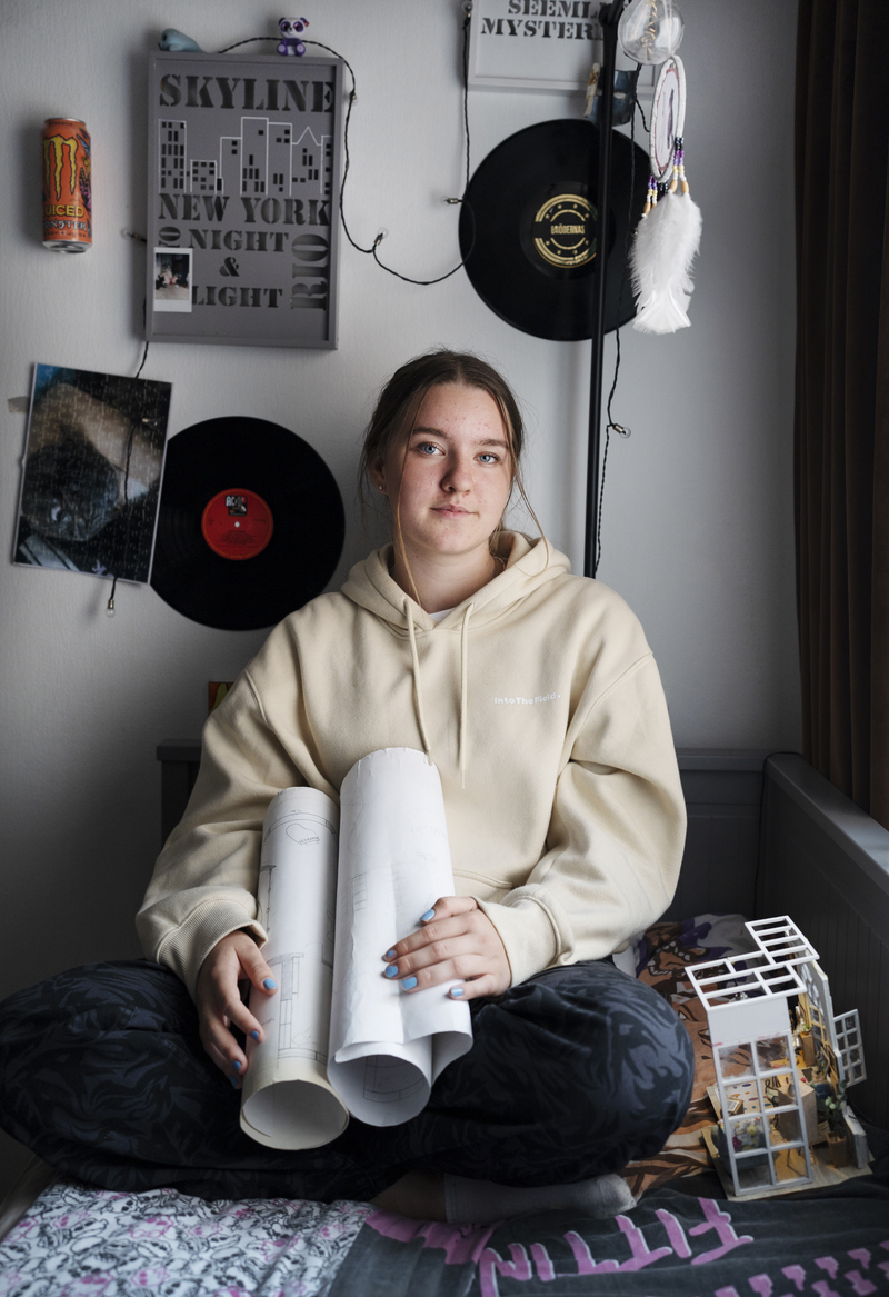Ung tjej sitter lutad mot en vägg, väggen är dekorerad med lp-skivor och hon har en hus-ritning i händerna
