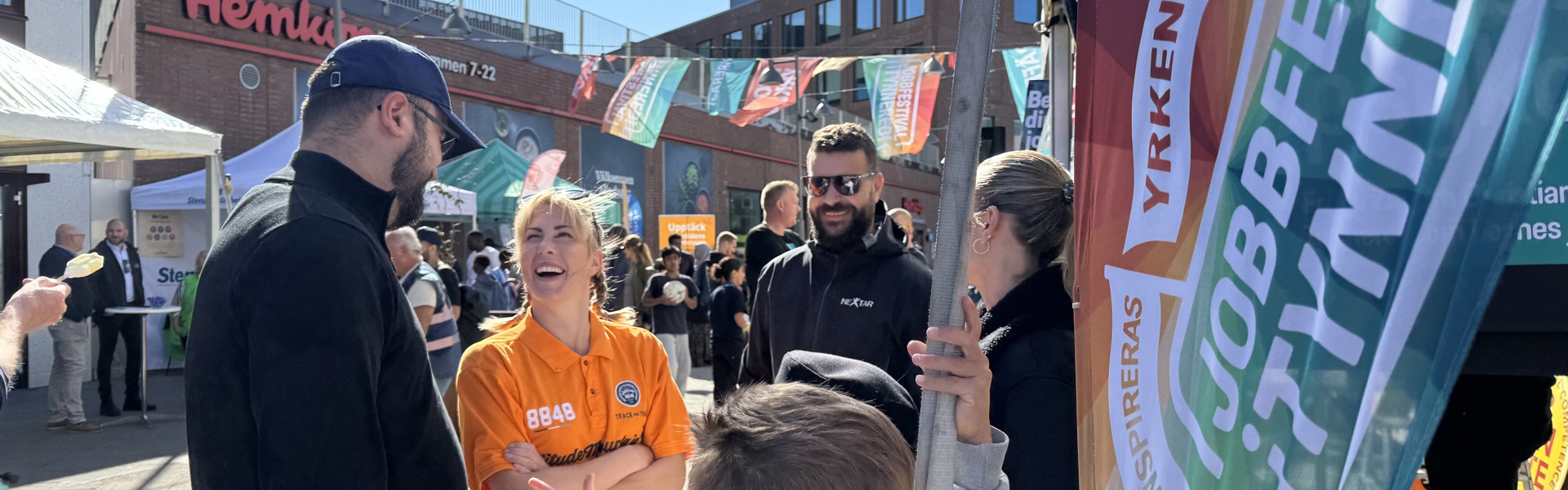 Människor möts och samtalar på Jobbfestivalen i Tynnered vid Opaltorget, med färgglada flaggor och aktiviteter i soligt väder.