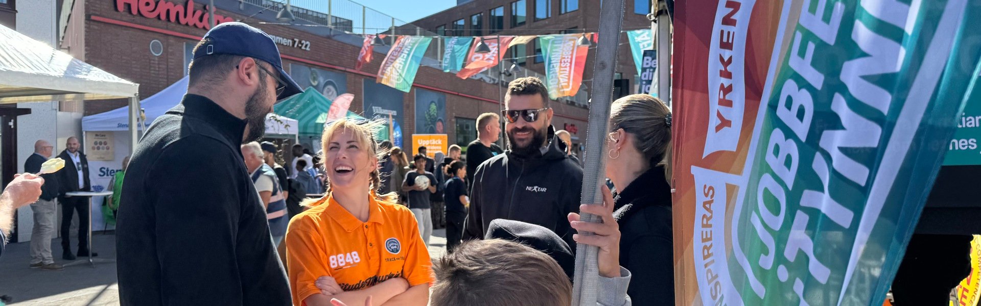 Människor möts och samtalar på Jobbfestivalen i Tynnered vid Opaltorget, med färgglada flaggor och aktiviteter i soligt väder.