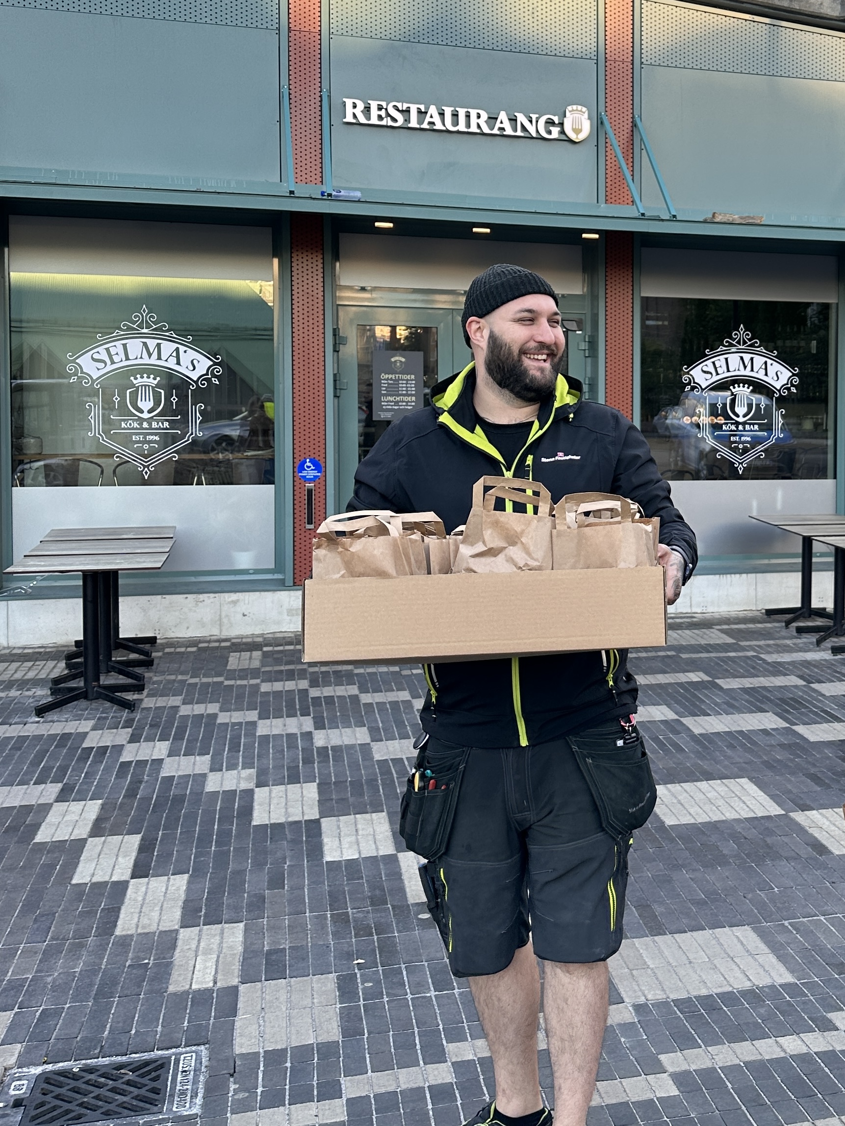 Person står utanför restaurangen Selma’s och ler medan hen bär en stor kartong fylld med flera bruna papperspåsar, på en stenlagd uteplats framför restaurangens glasdörrar.