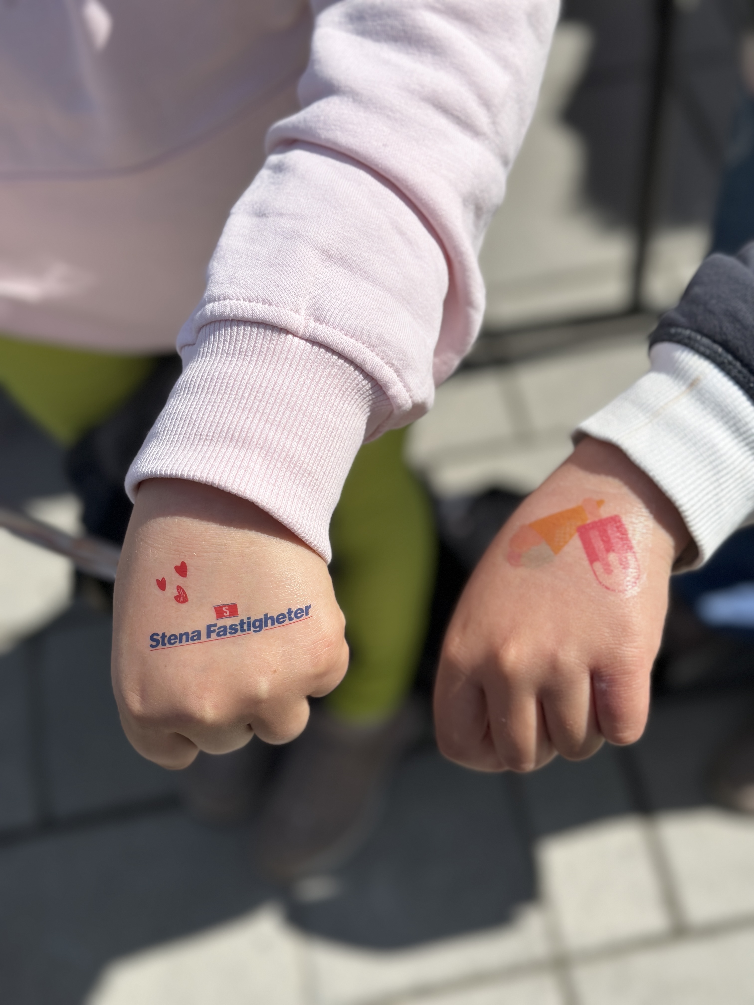 Två barns knutna händer i fokus utomhus, där den ena har en tillfällig tatuering med Stena Fastigheter‑logga och små hjärtan och den andra en färgglad bild, mot suddig bakgrund av markplattor och ben.