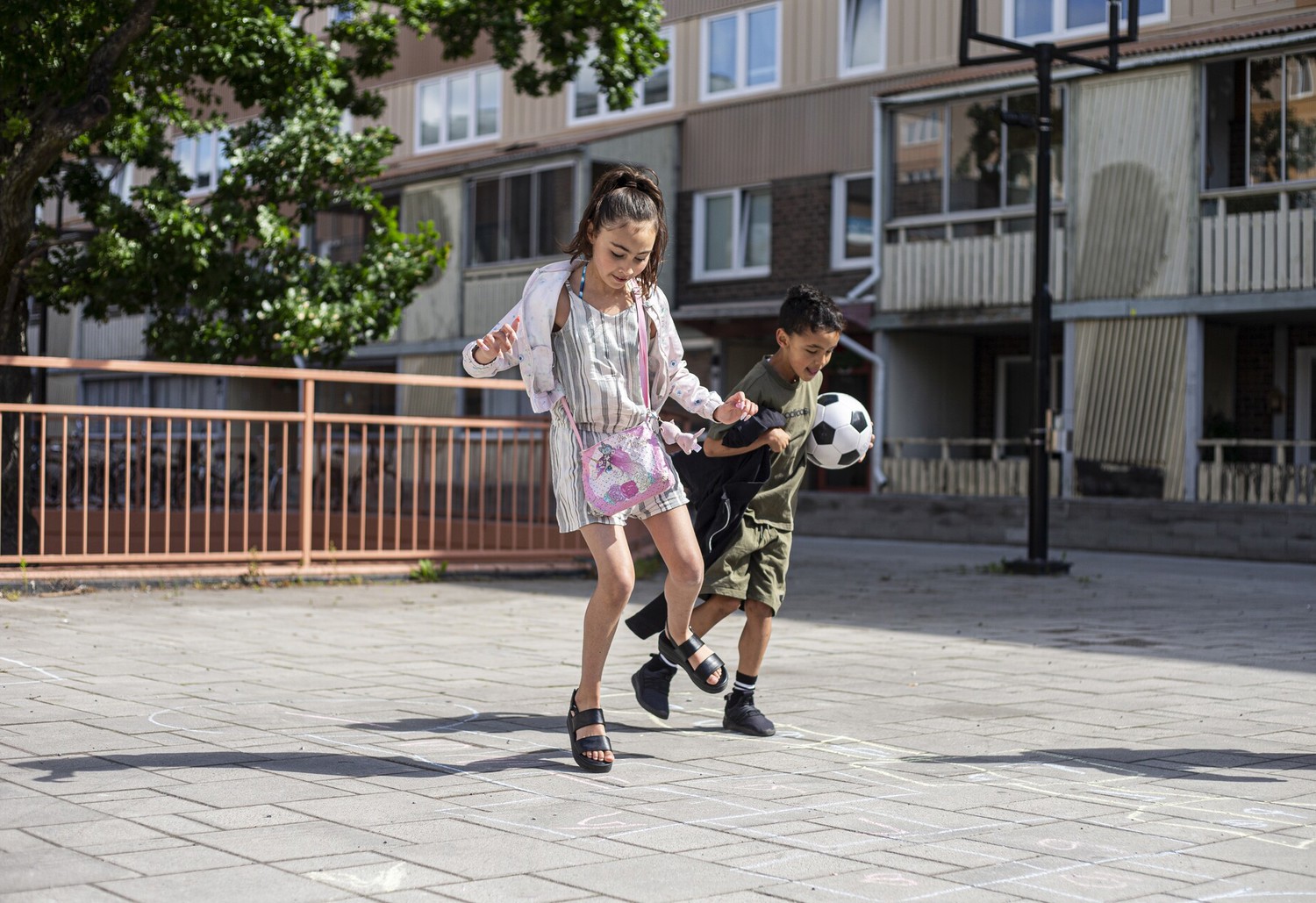 Två barn leker på en solig gård framför ett flerbostadshus; flickan hoppar på ett hopphagaritat med gatukritor medan pojken bredvid spelar fotbol.