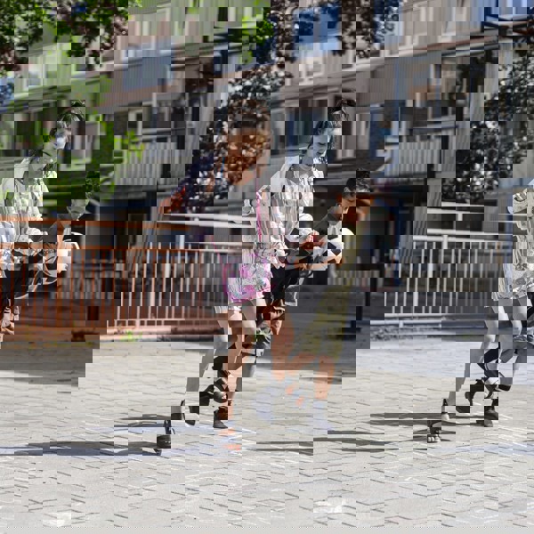 Två barn leker på en solig gård framför ett flerbostadshus; flickan hoppar på ett hopphagaritat med gatukritor medan pojken bredvid spelar fotbol.