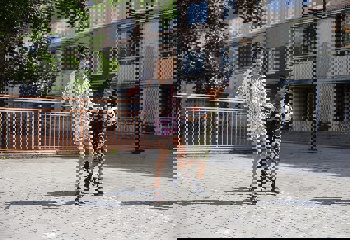 Två barn leker på en solig gård framför ett flerbostadshus; flickan hoppar på ett hopphagaritat med gatukritor medan pojken bredvid spelar fotbol.