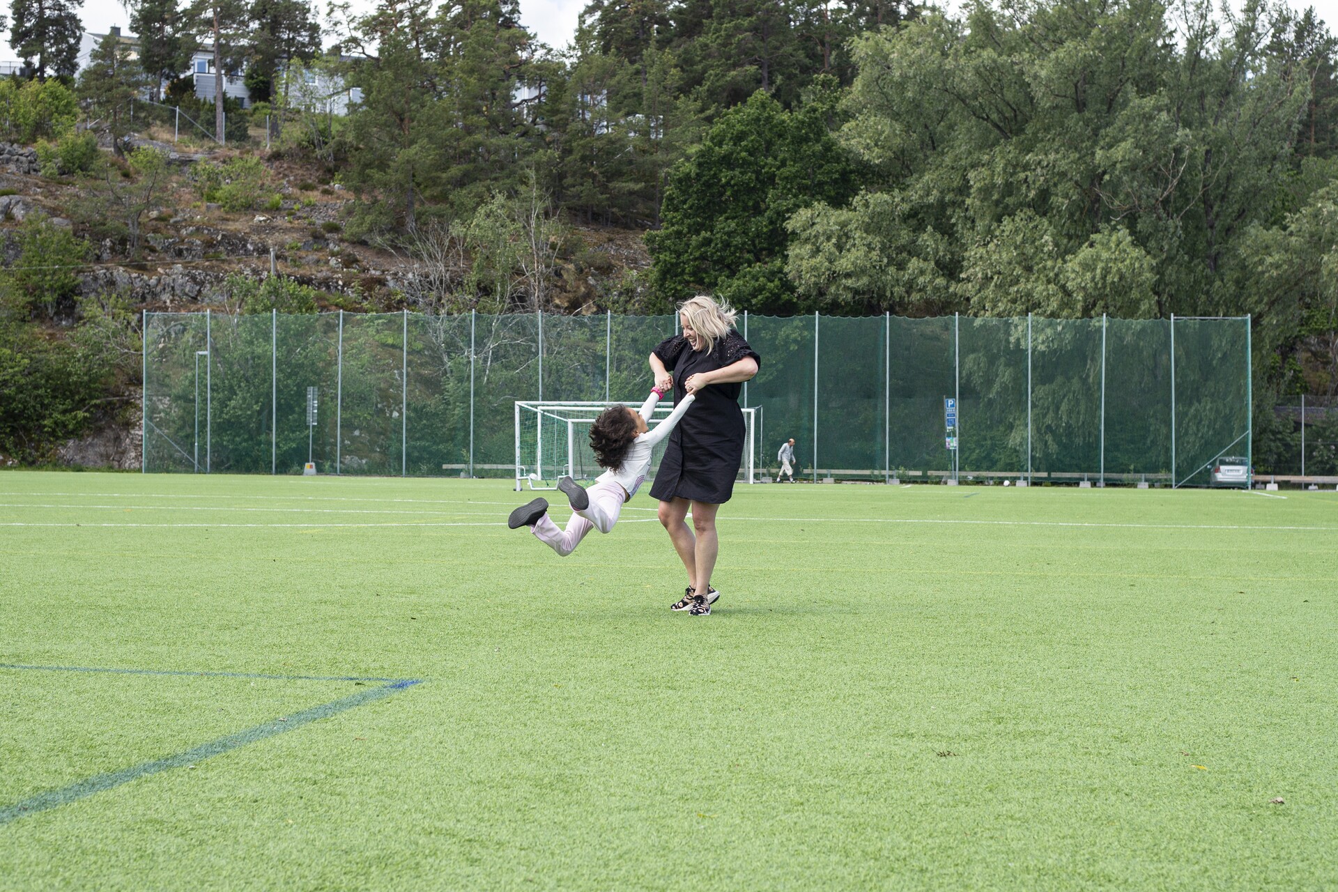 Kvinna i svart klänning snurrar runt ett barn som hänger i hennes händer på en stor grön fotbollsplan, med skogklädda berg och höga nätstaket i bakgrunden en mulen sommardag.