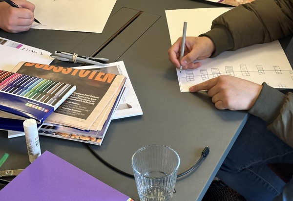 Två personer sitter vid ett bord och skissar på papper med pennor, omgivna av färgpennor, tidningar, sax, limstift, färgade papper och inspirationsbilder, vilket ger intryck av en kreativ workshop. Unga i Sao-jobb