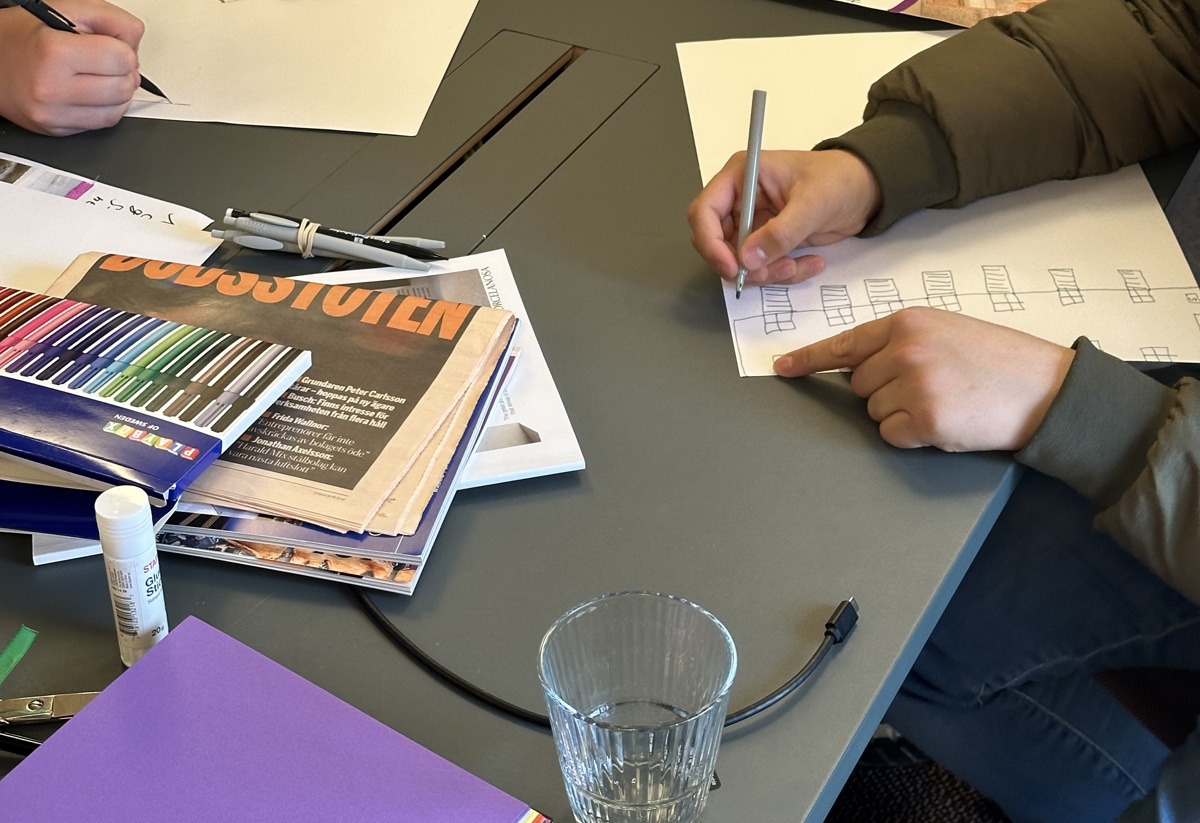 Två personer sitter vid ett bord och skissar på papper med pennor, omgivna av färgpennor, tidningar, sax, limstift, färgade papper och inspirationsbilder, vilket ger intryck av en kreativ workshop. Unga i Sao-jobb