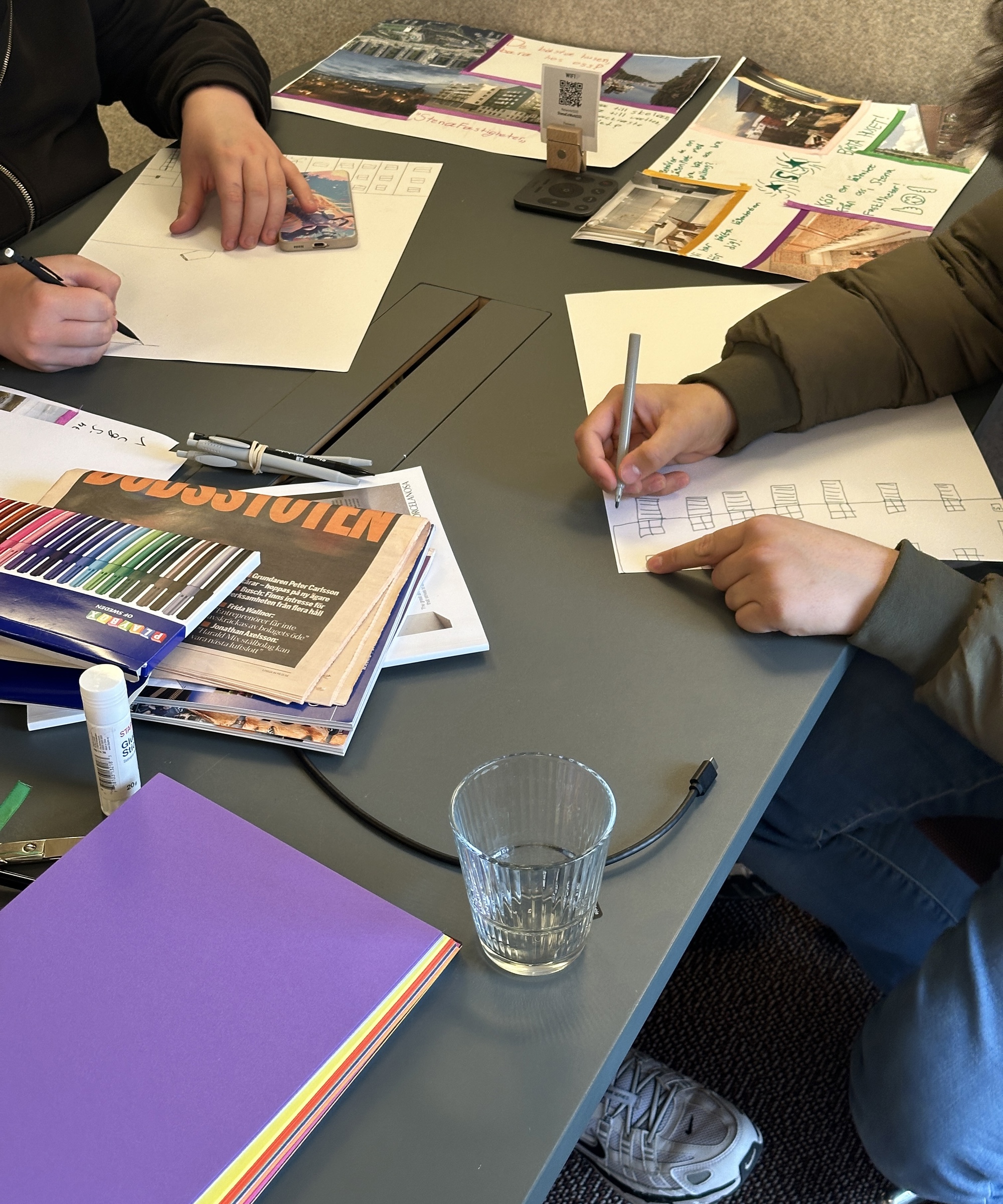 Två personer sitter vid ett bord och skissar på papper med pennor, omgivna av färgpennor, tidningar, sax, limstift, färgade papper och inspirationsbilder, vilket ger intryck av en kreativ workshop. Unga i Sao-jobb