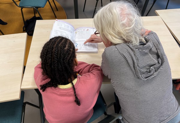 En äldre person med vitt hår och glasögon sitter bredvid ett barn med flätor vid ett bord i ett klassrum. De lutar sig båda över ett öppet arbetsblad med siffror och små illustrationer, medan den äldre personen pekar på något i boken. Runt dem syns tomma stolar och bord, vilket ger intryck av en lugn undervisnings- eller stunden för läxhjälp.