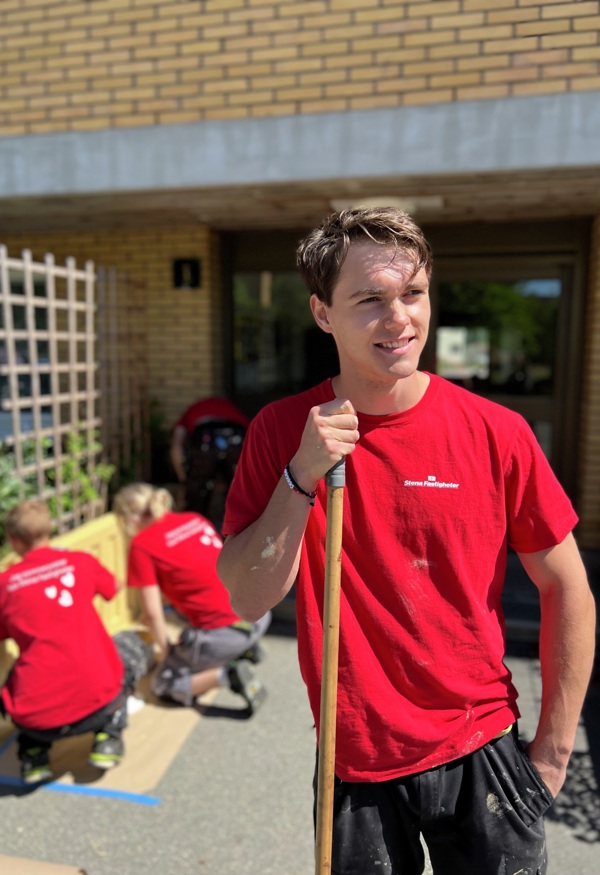 Ung person i röd Stena Fastigheter‑tröja står ute med ett träskaft i handen framför ett gult tegelhus, medan flera andra i likadana tröjor arbetar med planteringslådor på marken i bakgrunden.