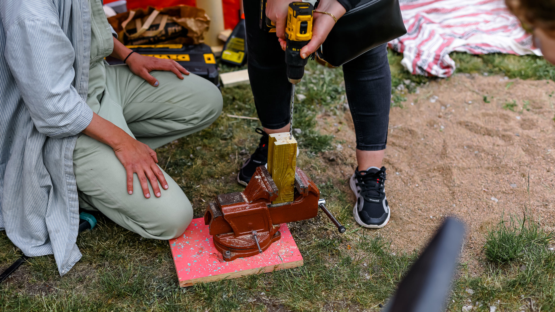 I mitten borrar en person ner i en träkloss, medan en annan person sitter på huk vid sidan. Marken består av en blandning av gräs och sand. Inga ansikten syns, endast kropparna.