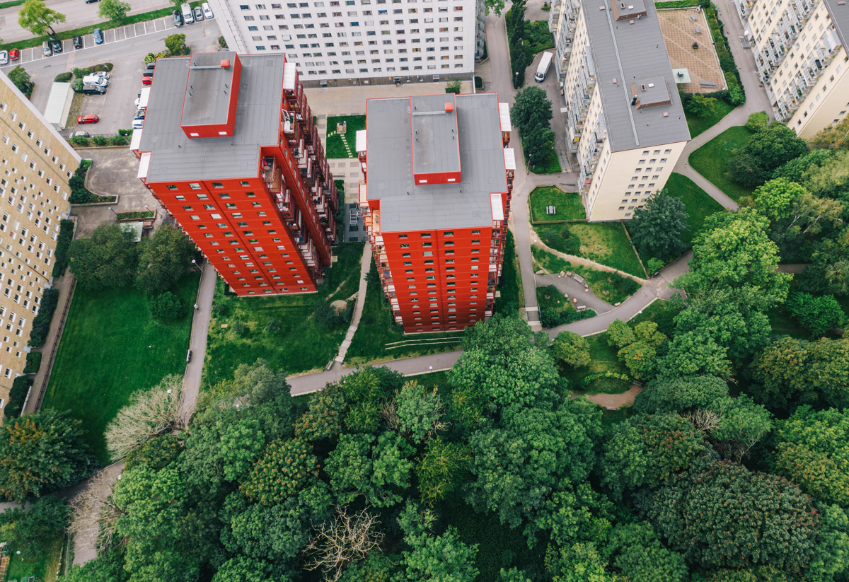 Drönarbild tagen från ovan på en stadsdel. På nedre halvan finns det träd och grönska samt en väg. På bildens övre halva visas sex höghus och en parkering med bilar. 