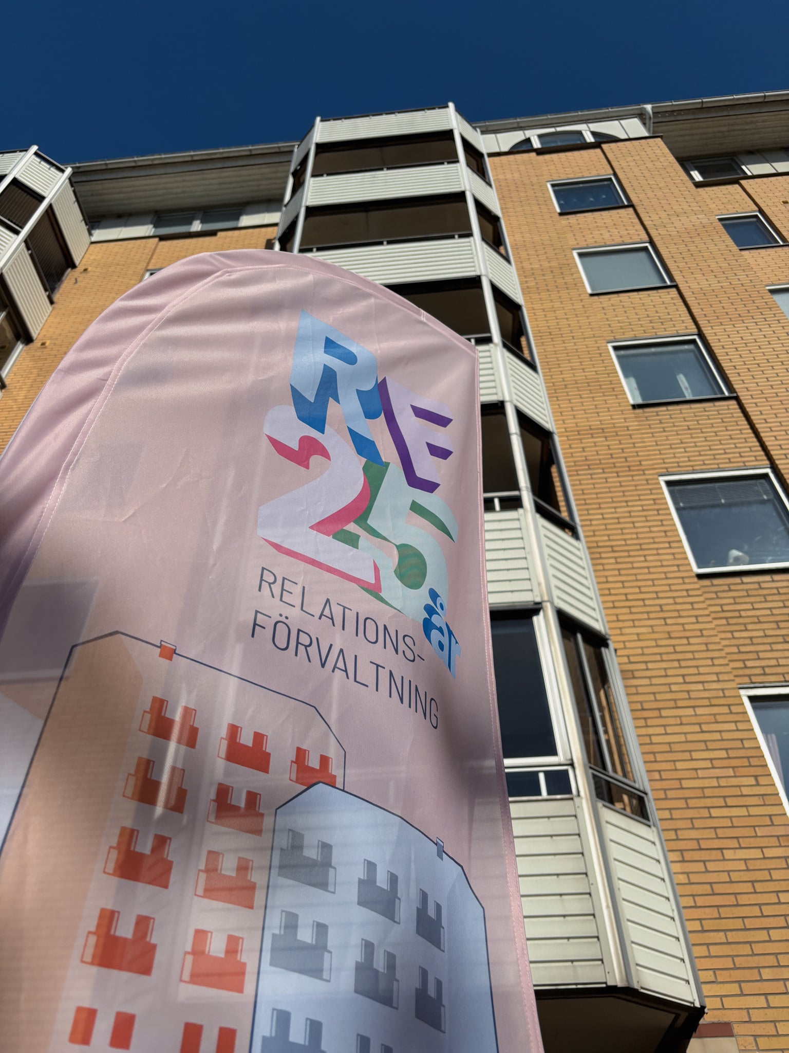  Vinklad närbild underifrån på en rosa flagga med texten ”RF 25 år – Relationsförvaltning” framför ett gulbrunt flerbostadshus mot klar blå himmel.