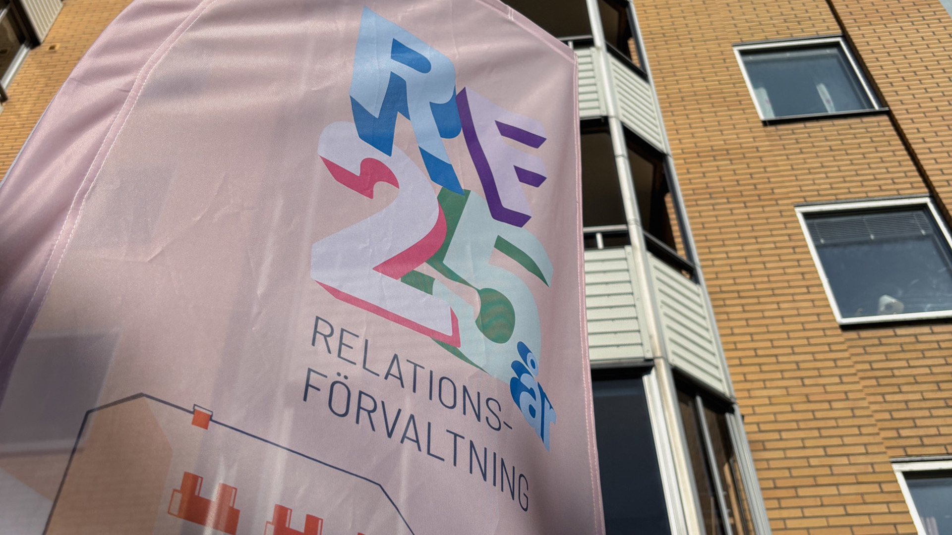 Done  Done Vinklad närbild underifrån på en rosa flagga med texten ”RF 25 år – Relationsförvaltning” framför ett gulbrunt flerbostadshus mot klar blå himmel.