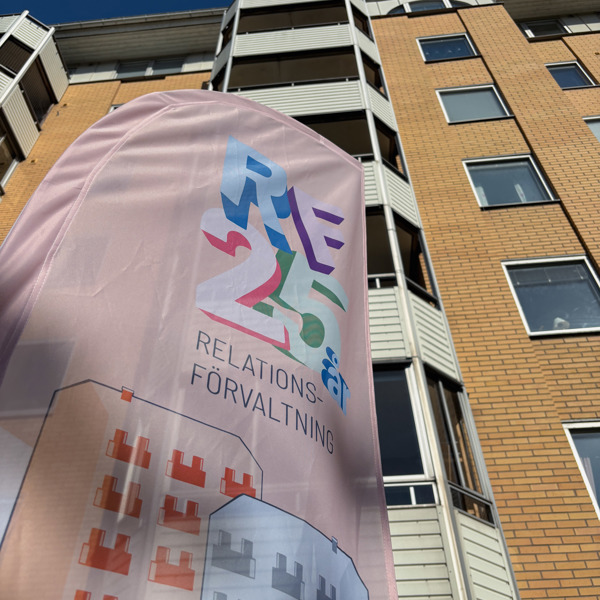  Vinklad närbild underifrån på en rosa flagga med texten ”RF 25 år – Relationsförvaltning” framför ett gulbrunt flerbostadshus mot klar blå himmel.