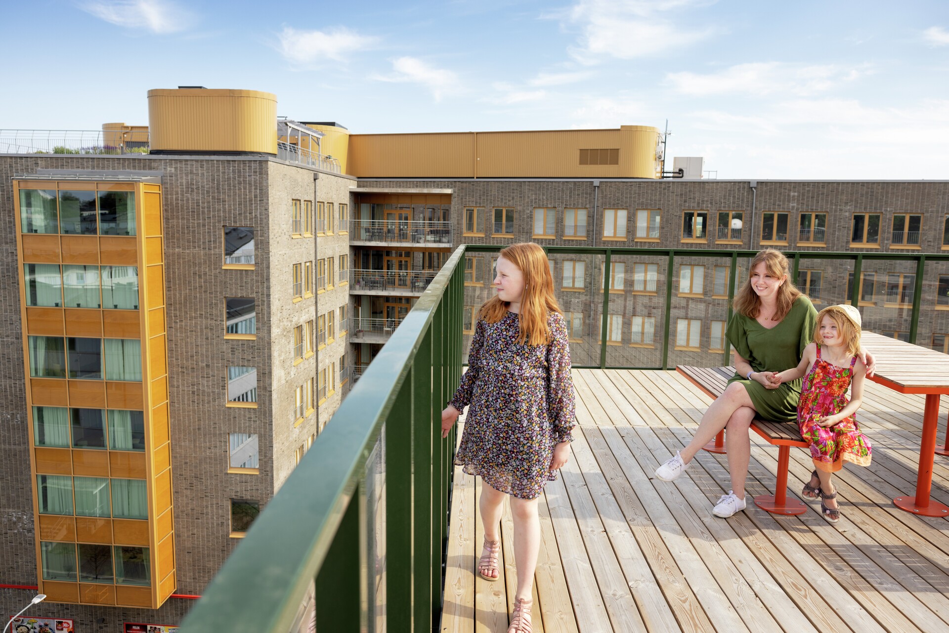 Tre personer på en takterrass en solig dag. Ett barn står nära balkongräcket och tittar ut. Bakom barnet sitter en kvinna och ett barn, båda ler mot barnet och håller varandra i handen.