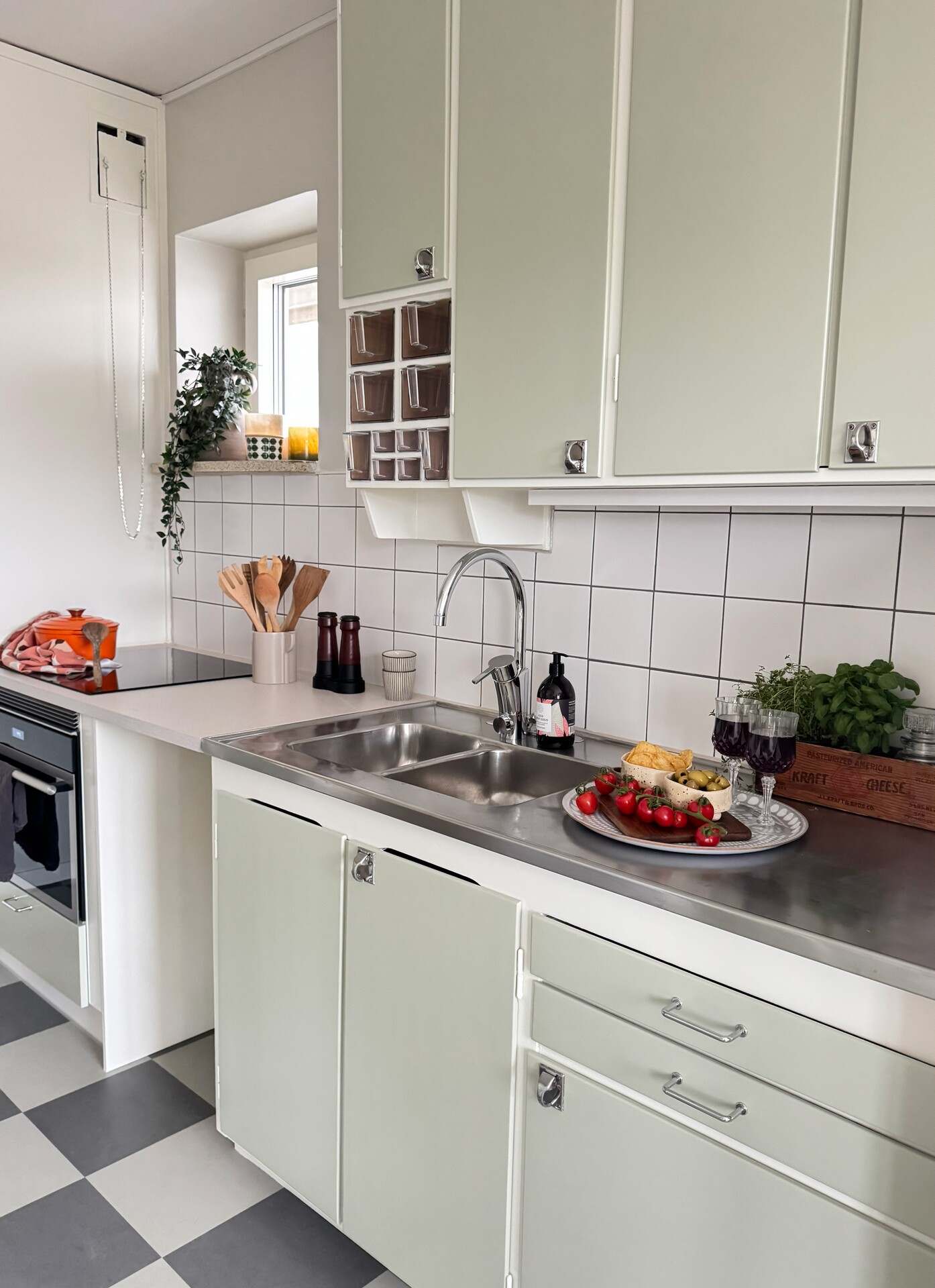 Ljust retroinspirerat kök med mintgröna skåpsluckor, vitt kakel och rostfri bänkskiva, där en dubbel diskho står i centrum. Till vänster syns en spishäll med orange gryta och träredskap i ett keramikställ, och vid fönstret står gröna växter och ljus. På bänken till höger om vasken finns en bricka med ost, kex, tomater och tre glas rött vin samt en trälåda med färska örter.