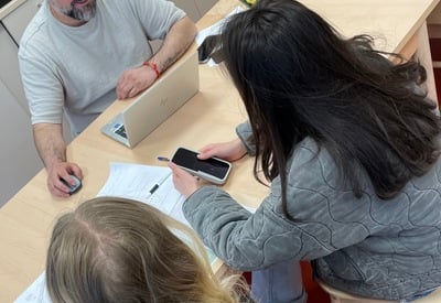 Tre personer sitter runt ett ljust träbord i ett kök eller pausrum. En man med skägg sitter framför en bärbar dator och tittar mot två ungdomar som sitter mittemot med papper och mobiltelefon på bordet. 