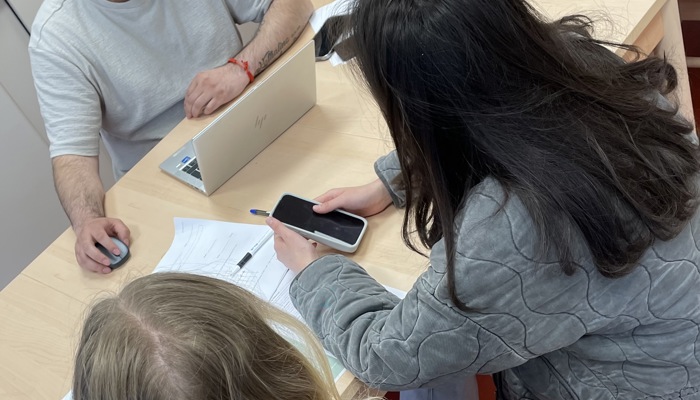 Tre personer sitter runt ett ljust träbord i ett kök eller pausrum. En man med skägg sitter framför en bärbar dator och tittar mot två ungdomar som sitter mittemot med papper och mobiltelefon på bordet. 