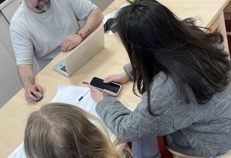 Tre personer sitter runt ett ljust träbord i ett kök eller pausrum. En man med skägg sitter framför en bärbar dator och tittar mot två ungdomar som sitter mittemot med papper och mobiltelefon på bordet. 