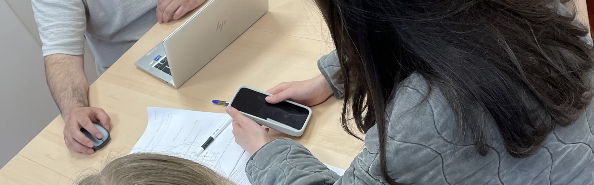 Tre personer sitter runt ett ljust träbord i ett kök eller pausrum. En man med skägg sitter framför en bärbar dator och tittar mot två ungdomar som sitter mittemot med papper och mobiltelefon på bordet. 