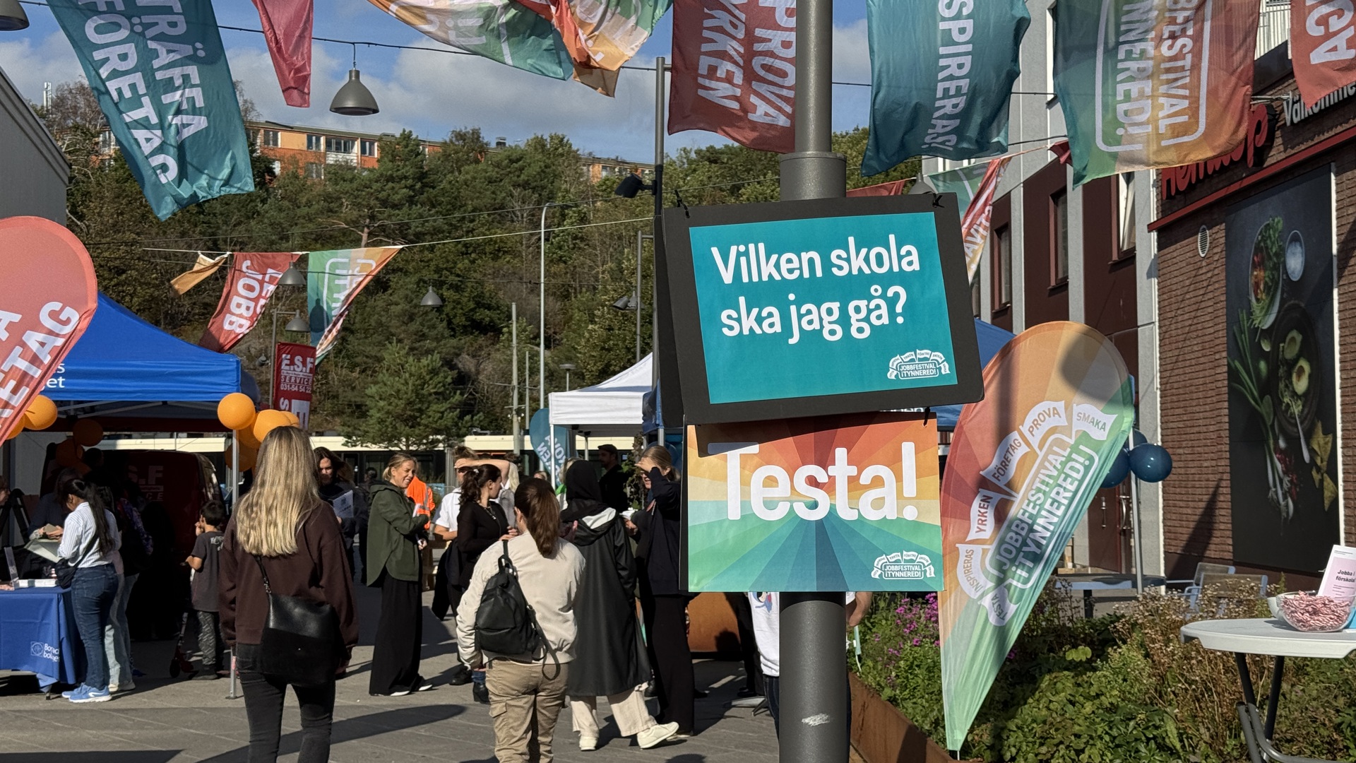 Utomhusmässa på ett torg med många besökare, färgglada tält och flaggor. I mitten sitter en skylt på en lyktstolpe med texten ”Vilken skola ska jag gå? Testa!”, och runtom syns fler banderoller med ord om att träffa företag och prova yrken. I bakgrunden skymtar träd och bostadshu