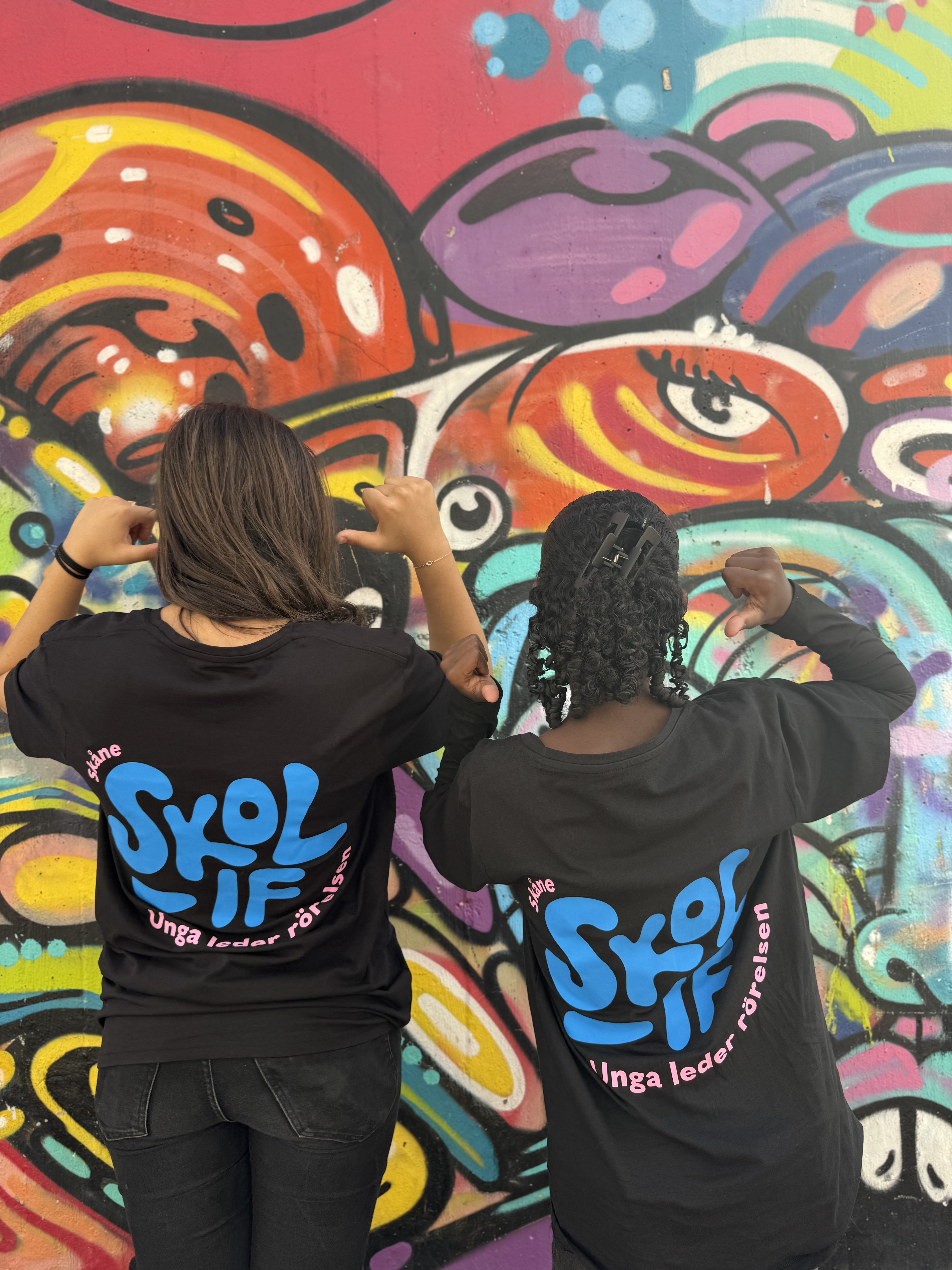 Två barn med ryggen mot kameran står framför en färgstark graffitivägg och pekar med tummarna mot sina svarta T‑tröjor med blå och rosa texten ”Skol IF – Unga leder rörelsen” på ryggen.