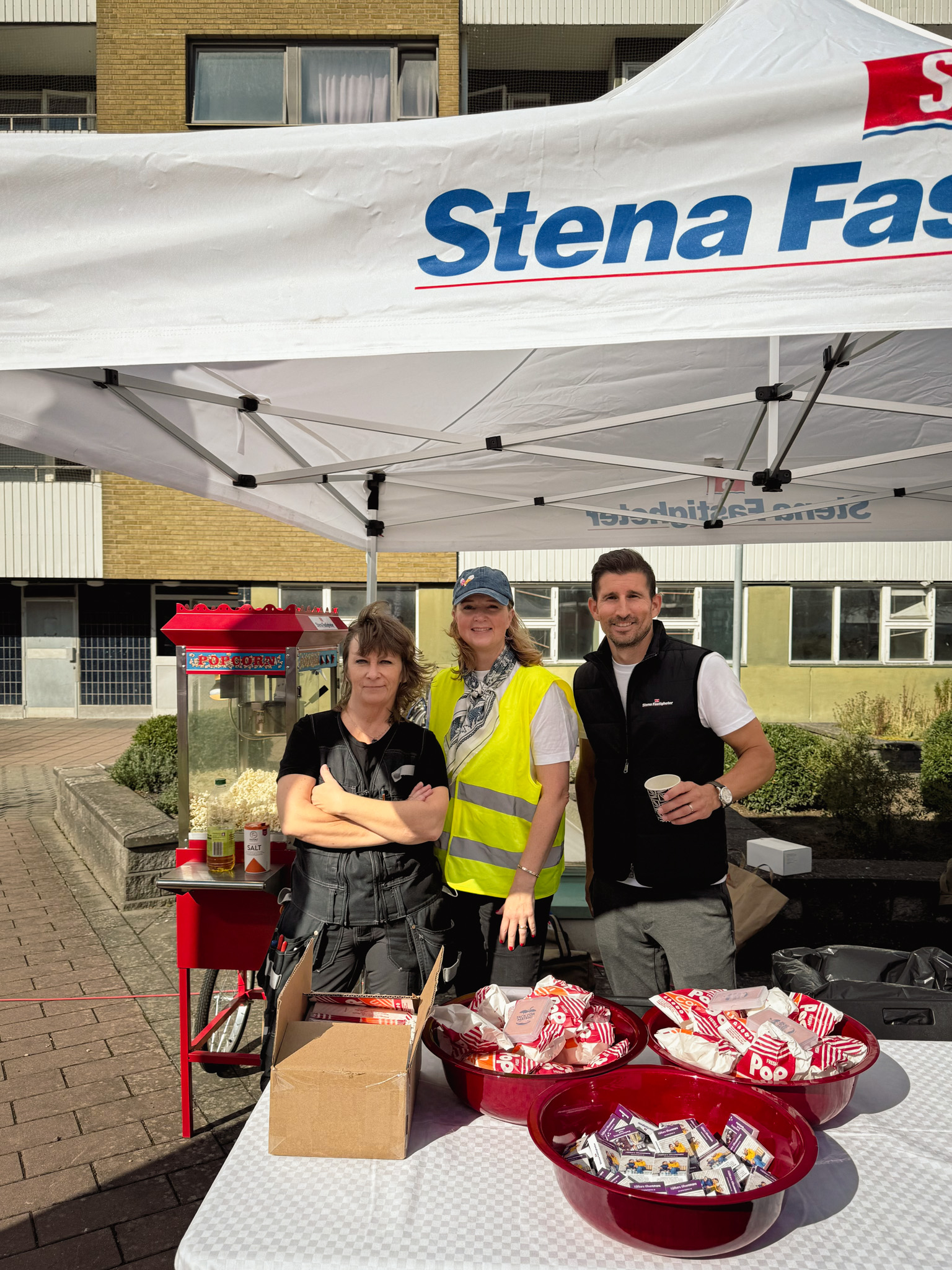 Tre personer står under ett vitt tält med Stena Fastigheter‑logga på ett gårdstorg. Framför dem står en popcornmaskin och ett bord med stora röda skålar och kartonger fyllda med popcornpåsar och godis.