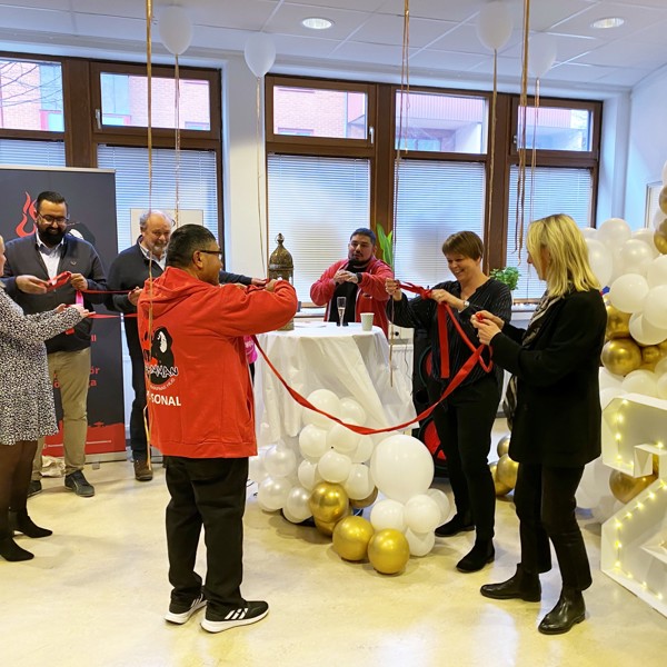 En grupp människor deltar i en bandklippningsceremoni inomhus. Rummet är dekorerat med vita och guldiga ballonger. En person i röd jacka klipper bandet, medan andra ser på och ler. Stämningen är festlig och högtidlig.