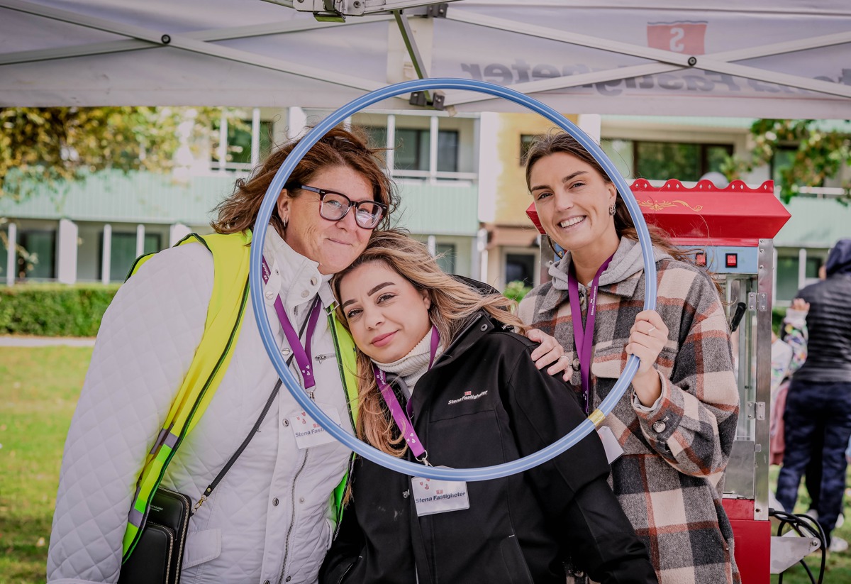 Tre personer står tillsammans under ett tält och ler genom en rockring. De bär namnskyltar och verkar delta i ett evenemang. I bakgrunden syns en grön gräsmatta och en byggnad. Stämningen är glad och gemytlig.