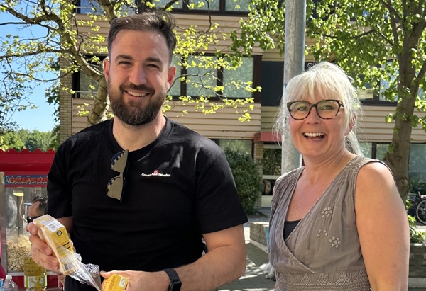 Två personer står utomhus och ler mot kameran. De håller i snacks och drycker. Solen skiner och träden i bakgrunden är gröna, vilket ger en somrig känsla. En byggnad syns i bakgrunden.