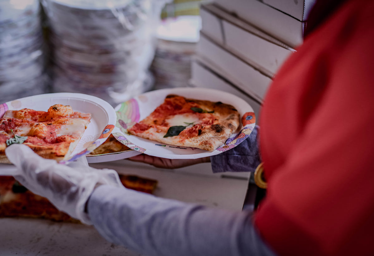 En person i röd tröja och handskar serverar pizzabitar på papptallrikar. I bakgrunden syns staplade pizzakartonger. Bilden ger en känsla av matservering vid ett evenemang eller en fest.