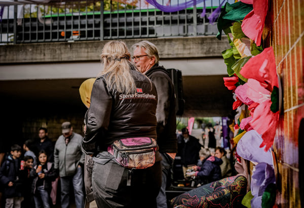 En grupp människor står utomhus vid ett evenemang. En person bär en jacka med text på ryggen. Färgglada dekorationer syns i förgrunden, och fler personer är samlade i bakgrunden. Stämningen verkar livlig och social.
