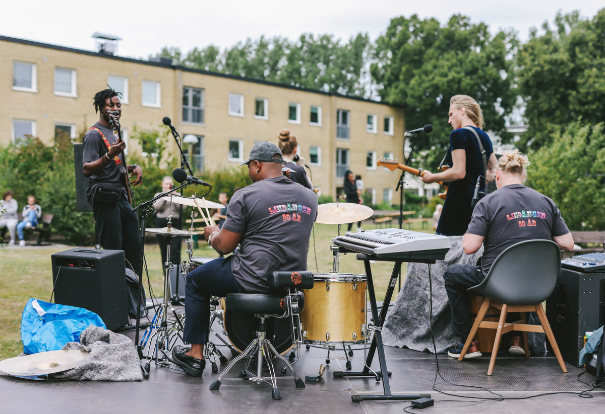 Ett band spelar musik utomhus på en scen. Medlemmarna spelar olika instrument som trummor, gitarr och keyboard. I bakgrunden syns en byggnad och grönska. Publiken sitter utspridd på gräset och lyssnar. Stämningen är avslappnad och festlig.