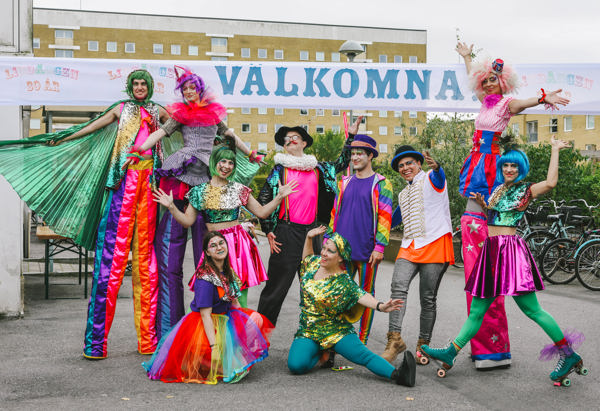 En grupp färgglatt klädda personer poserar framför en skylt med texten "VÄLKOMNA". Några av dem står på styltor, och deras kläder är i livliga färger. Bakom dem syns en byggnad. Stämningen är festlig och inbjudande.