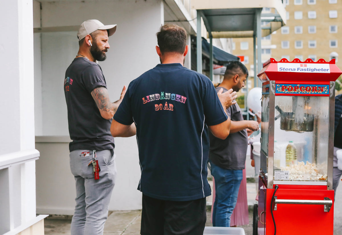 Flera personer står utomhus vid en popcornmaskin. De är klädda i t-shirts och verkar samtala. I bakgrunden syns en byggnad. Stämningen är avslappnad och social, som vid ett utomhusevenemang.