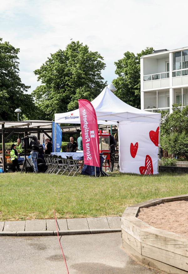 Ett evenemangstält står uppställt på en gräsmatta nära en lekplats. Tältet är dekorerat med hjärtan och omges av färgglada flaggor. I bakgrunden syns en modern byggnad och grönskande träd. Stämningen är inbjudande och somrig.