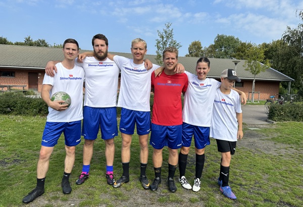 Sex personer står på en gräsplan i fotbollskläder. De har vita och röda tröjor och blå shorts. En av dem håller en fotboll. Gruppen ser glad ut och omfamnar varandra, med en klar himmel i bakgrunden. Stämningen är kamratlig och sportig.