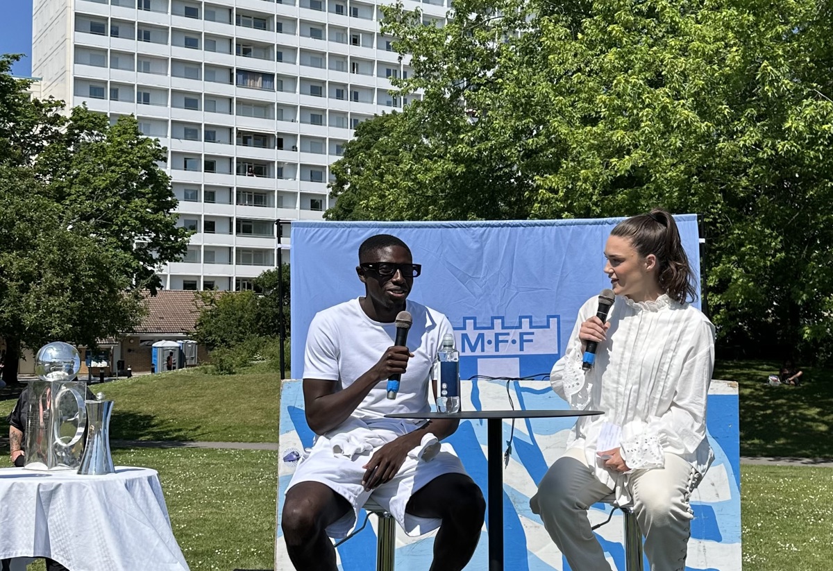 Två personer sitter på höga stolar utomhus och håller mikrofoner. Bakom dem finns en skylt med MFF-logotypen. En av dem är klädd i vitt, och bredvid dem står ett bord med en pokal. I bakgrunden syns en hög byggnad och grönska. Stämningen är professionell och somrig.