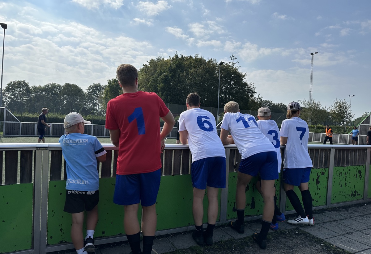 En grupp personer i fotbollskläder står vid ett staket och tittar på en match. De har nummer på sina tröjor och bär blå shorts. Solen skiner, och himlen är delvis molnig. Omgivningen är grönskande och sportig.
