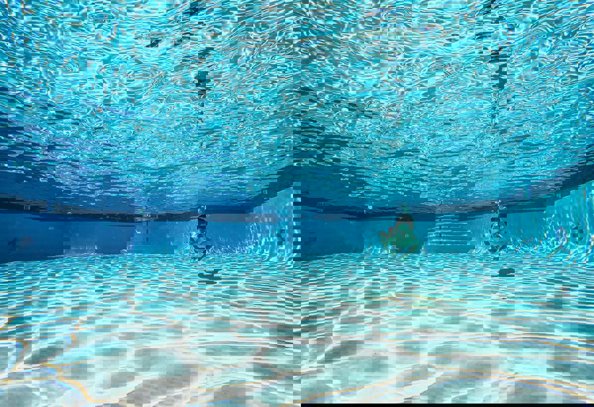 En undervattensbild av en pool med klart, blått vatten. Solens strålar skapar ett vackert mönster på botten. En person simmar under vattnet, vilket ger en känsla av lugn och frihet.