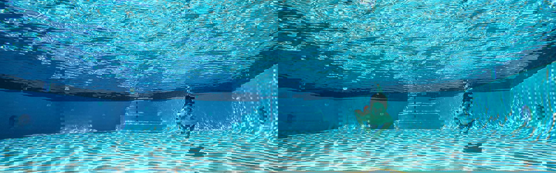 En undervattensbild av en pool med klart, blått vatten. Solens strålar skapar ett vackert mönster på botten. En person simmar under vattnet, vilket ger en känsla av lugn och frihet.
