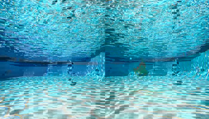 En undervattensbild av en pool med klart, blått vatten. Solens strålar skapar ett vackert mönster på botten. En person simmar under vattnet, vilket ger en känsla av lugn och frihet.
