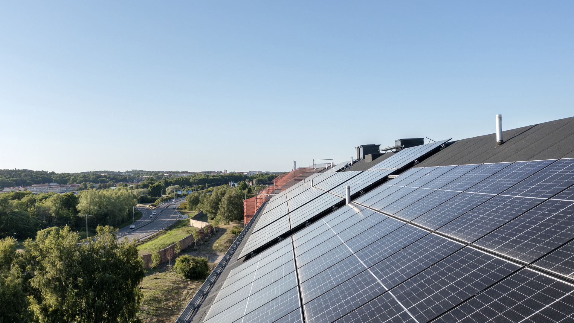 Solpaneler täcker ett snedtak på en byggnad i förgrunden, med grönskande träd, en väg med bilar och ett stadslandskap i bakgrunden under klar blå himmel.