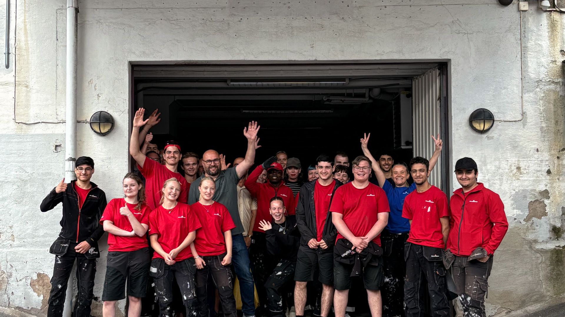En grupp personer i arbetskläder, många i röda tröjor, står samlade framför en öppen garageport. Flera ler och vinkar mot kameran, vilket ger en glad och gemensam lagkänsla.
