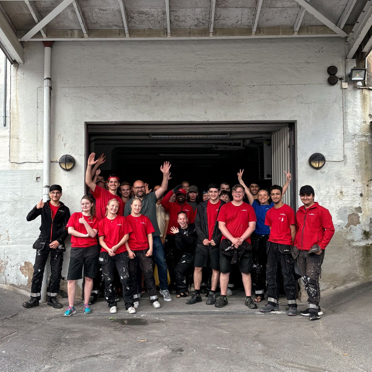 En grupp personer i arbetskläder, många i röda tröjor, står samlade framför en öppen garageport. Flera ler och vinkar mot kameran, vilket ger en glad och gemensam lagkänsla.