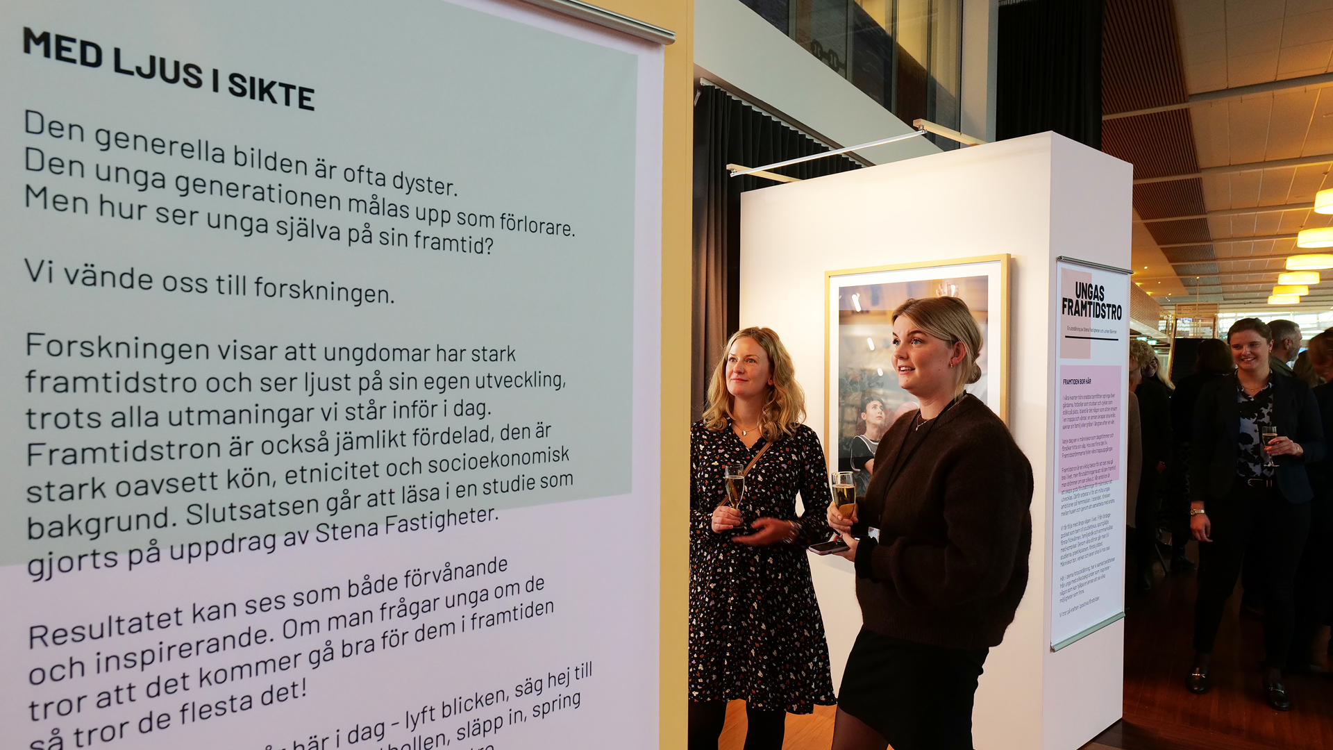 Till vänster syns en ljusgrön och ljusrosa affisch på väggen med rubriken ´Med ljus i sikte´ och en text som handlar om att vetenskap bevisar att ungdomar har framtidstro. I bakgrunden syns personer som tar del av utställningen ´Ungas framtidstro´ i Göteborg.  