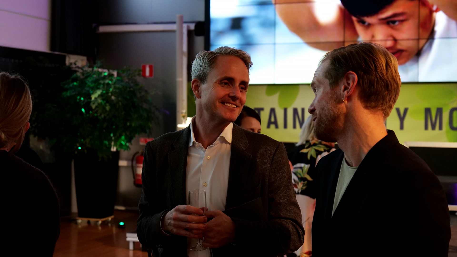 Två män iklädda kavaj kollar mot varandra och ser glada ut. I bakgrunden syns en scen och en stor skärm ovanför. 