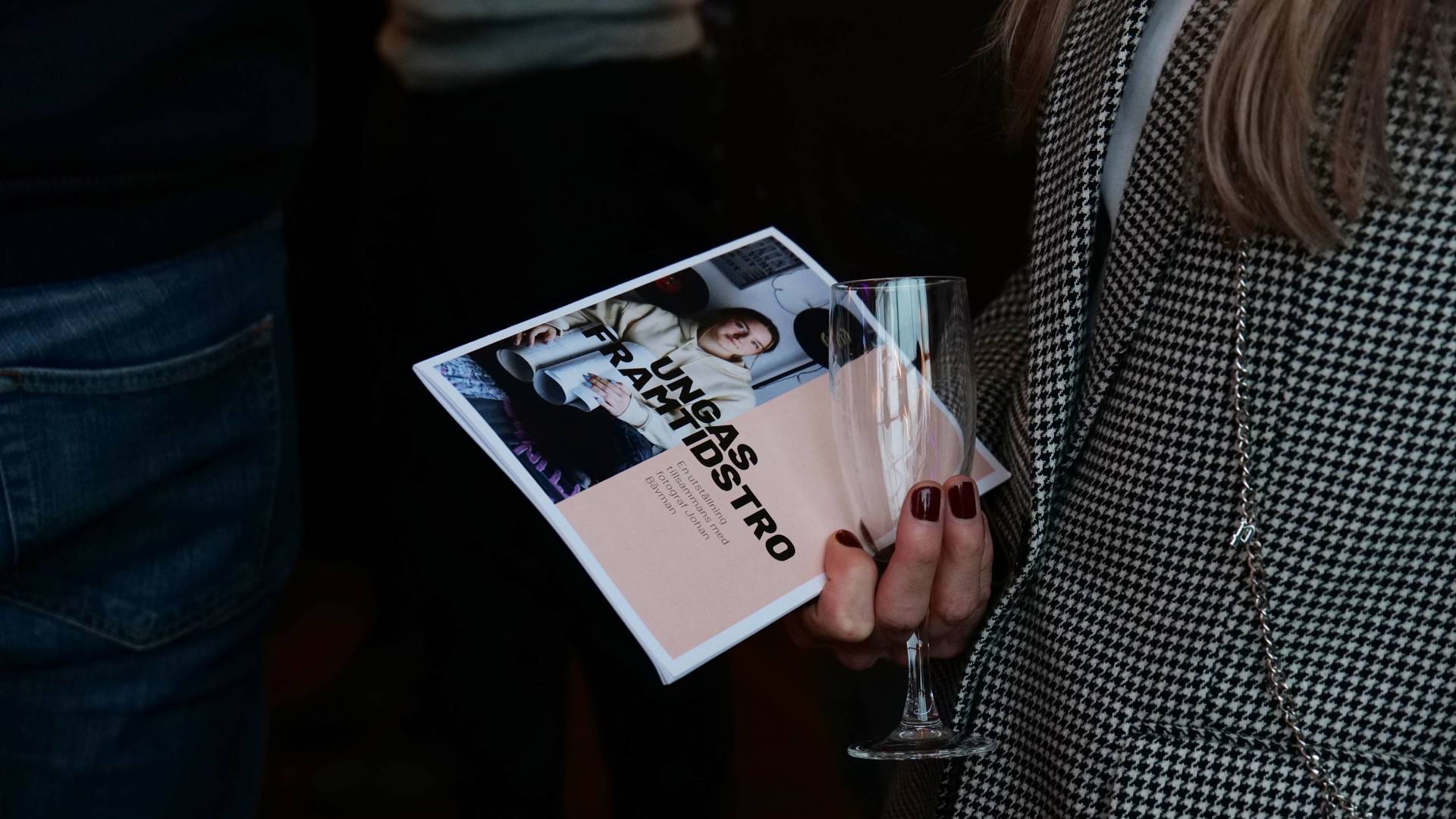 En kvinnas överkropp utan ansikte, iklädd en rutig kavaj. Hon håller ett champagneglas och en broschyr med texten ´ungas framtidstro´ i handen. 